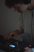 A person wearing headphones is using an electronic music production device. The focus is on their hands interacting with the equipment, surrounded by small, colorful lights on the device.