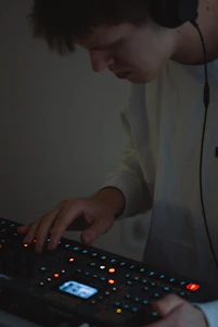 A person wearing headphones is using an electronic music production device. The focus is on their hands interacting with the equipment, surrounded by small, colorful lights on the device.