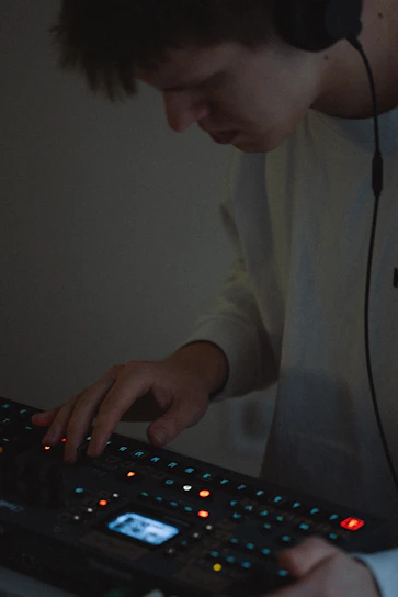 A person wearing headphones is using an electronic music production device. The focus is on their hands interacting with the equipment, surrounded by small, colorful lights on the device.