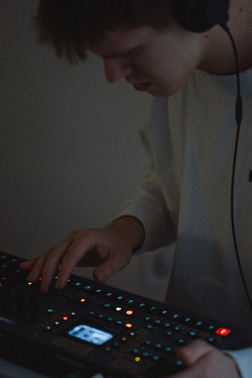 A person wearing headphones is using an electronic music production device. The focus is on their hands interacting with the equipment, surrounded by small, colorful lights on the device.