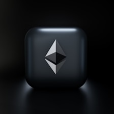 A sleek, modern design featuring a three-dimensional geometric shape resembling the Ethereum logo, centered on a glossy dark gray square with subtle lighting effects. The background fades into a deep black, highlighting the central emblem.