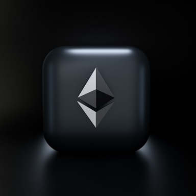 A sleek, modern design featuring a three-dimensional geometric shape resembling the Ethereum logo, centered on a glossy dark gray square with subtle lighting effects. The background fades into a deep black, highlighting the central emblem.