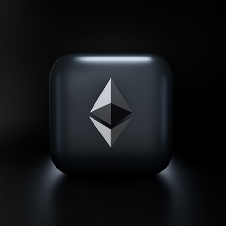 A sleek, modern design featuring a three-dimensional geometric shape resembling the Ethereum logo, centered on a glossy dark gray square with subtle lighting effects. The background fades into a deep black, highlighting the central emblem.