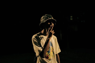 A person wearing a hat is partially illuminated against a dark background, holding something in front of their face. The t-shirt features a colorful graphic design with cartoon characters.