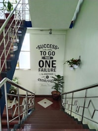 A stairwell features motivational text on the wall reading 'Success is the ability to go from one failure to another with no loss of enthusiasm - Winston Churchill'. The space is well-lit with natural light coming from a window. Several potted plants are placed around, enhancing the calm atmosphere. The staircase is adorned with metal railings.