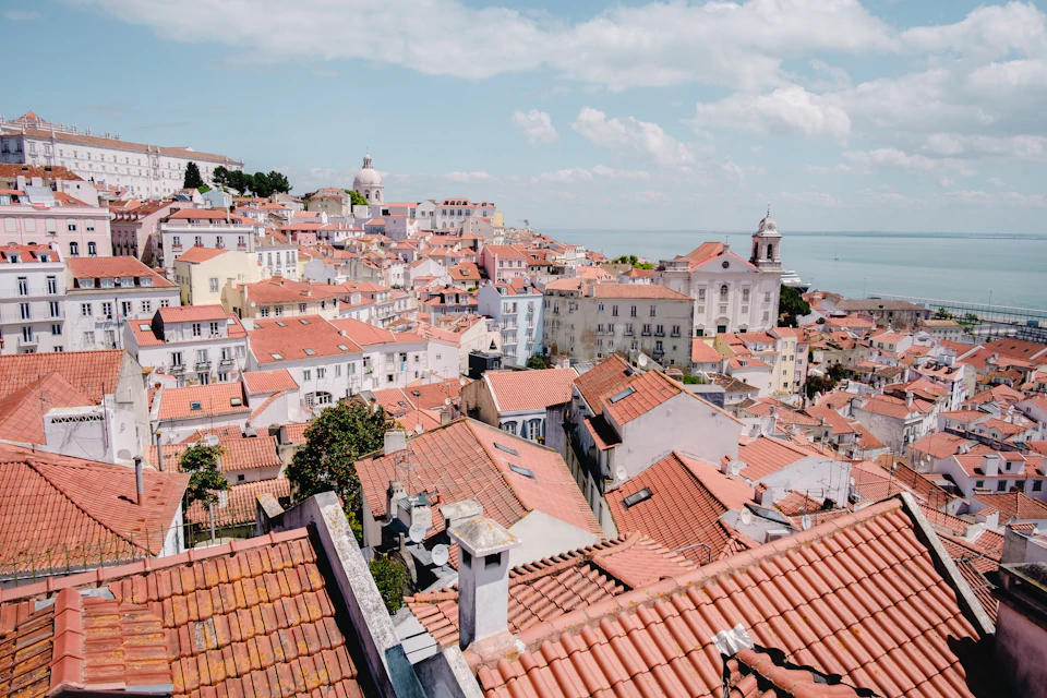 Lisbon's terracotta-red rooftops cascading down the hills of Alfama district under September's golden afternoon light, with the Tagus river visible in the distance
