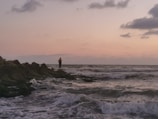 A traveler standing on a rocky shore watching waves crash at sunset.