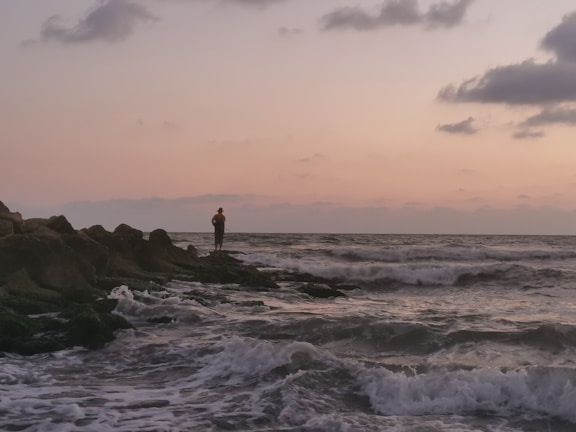 A traveler standing on a rocky shore watching waves crash at sunset.