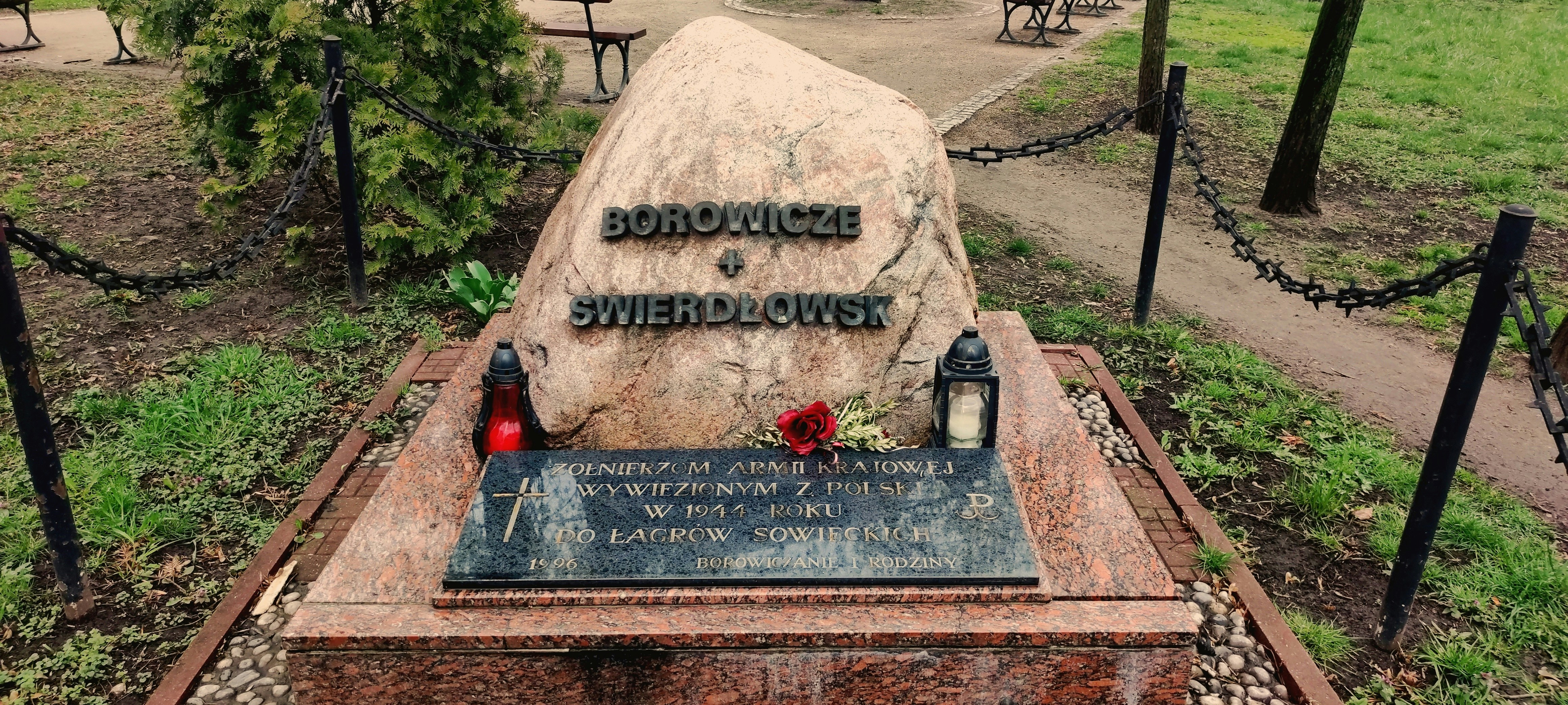A memorial stone with inscriptions is placed in a park setting. The stone is accompanied by two lanterns, one red and the other white, and a single red rose lies on the stone. The area is enclosed by a low chain-link fence with surrounding greenery and trees.