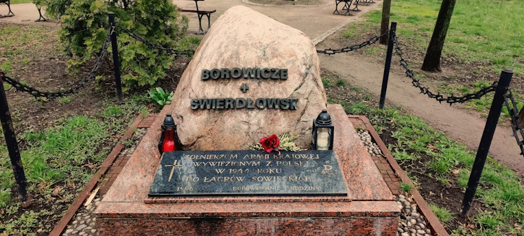 A memorial stone with inscriptions is placed in a park setting. The stone is accompanied by two lanterns, one red and the other white, and a single red rose lies on the stone. The area is enclosed by a low chain-link fence with surrounding greenery and trees.