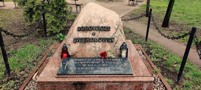 A memorial stone with inscriptions is placed in a park setting. The stone is accompanied by two lanterns, one red and the other white, and a single red rose lies on the stone. The area is enclosed by a low chain-link fence with surrounding greenery and trees.