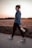 A person is walking on a paved path, holding a white water bottle. The individual is wearing athletic clothing, including a blue long-sleeve top, black leggings, and light-colored running shoes. The backdrop features an open field with short grass and a clear sky during sunset, adding a warm glow to the scene.