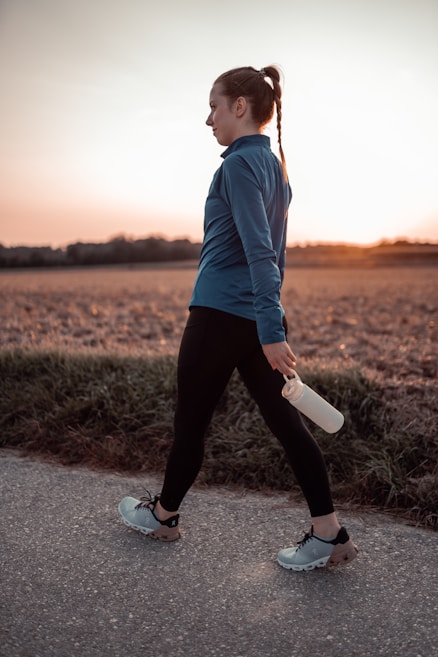 A person is walking on a paved path, holding a white water bottle. The individual is wearing athletic clothing, including a blue long-sleeve top, black leggings, and light-colored running shoes. The backdrop features an open field with short grass and a clear sky during sunset, adding a warm glow to the scene.