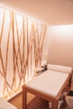 A minimalist spa room featuring a comfortable massage table with white sheets. The bamboo stalks attached vertically to the wall create an aesthetic, natural ambiance. Soft lighting enhances the calm and relaxing atmosphere, and a small stand with essential items is placed next to the table.