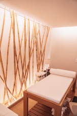 A minimalist spa room featuring a comfortable massage table with white sheets. The bamboo stalks attached vertically to the wall create an aesthetic, natural ambiance. Soft lighting enhances the calm and relaxing atmosphere, and a small stand with essential items is placed next to the table.