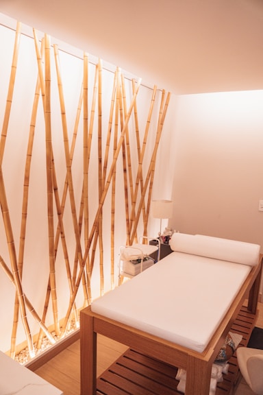 A minimalist spa room featuring a comfortable massage table with white sheets. The bamboo stalks attached vertically to the wall create an aesthetic, natural ambiance. Soft lighting enhances the calm and relaxing atmosphere, and a small stand with essential items is placed next to the table.