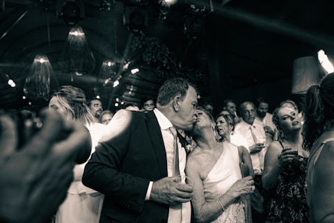 A group of people are gathered at a celebration or event, dressed formally. A couple in the foreground shares a kiss amidst the crowd, which includes people holding drinks, smiling, and engaged in conversation.