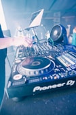A DJ setup with a turntable and various controls is prominently featured. A hand is adjusting one of the knobs on the mixer, with a laptop open in the background displaying the setup for a music performance. The environment suggests a lively and technical setting with electronic equipment.