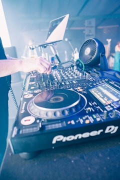 A DJ setup with a turntable and various controls is prominently featured. A hand is adjusting one of the knobs on the mixer, with a laptop open in the background displaying the setup for a music performance. The environment suggests a lively and technical setting with electronic equipment.