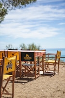 A rustic outdoor seating area with wooden director-style chairs surrounding a table draped with a red and white checkered tablecloth. The chairs feature yellow fabric backs with 'Moritz Barcelona' branding. In the background, there is a view of the sea under a clear blue sky with scattered clouds. The ground is covered with dirt and small pebbles.