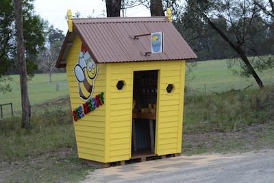 A small yellow wooden hut with a brown roof situated in a grassy area surrounded by trees. The hut features a cartoon bee design and colorful letters spelling a message on the side. Inside, jars and containers are visible on shelves. The surrounding area appears rural and natural.