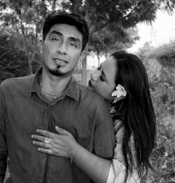 A man and woman are standing closely together in an outdoor setting with trees in the background. The woman is leaning her head affectionately on the man's shoulder, wearing a flower in her hair and jewelry on her fingers. The image is primarily in black and white with a slight green tint.