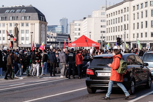 A large group of people gathered on a city street, some holding flags and signs. The setting features multiple buildings and a tall statue, and there's a red canopy tent in the background. Cars are seen parked nearby, and the crowd includes individuals in jackets and hoodies.