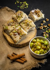 A rustic presentation featuring pieces of dessert topped with chopped nuts and garnished with green pistachios, arranged on a wooden slab. Beside it is a small white bowl filled with green olives. The backdrop includes a textured burlap surface and some cinnamon sticks, creating a warm aesthetic.