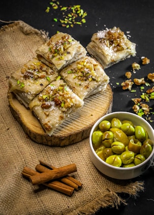 A rustic presentation featuring pieces of dessert topped with chopped nuts and garnished with green pistachios, arranged on a wooden slab. Beside it is a small white bowl filled with green olives. The backdrop includes a textured burlap surface and some cinnamon sticks, creating a warm aesthetic.