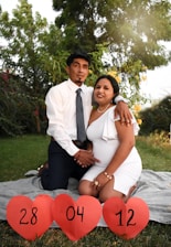A couple is sitting on a blanket in a garden setting. The woman is wearing a white dress and the man is wearing a white shirt with a tie. They are holding hands and smiling. In front of them, three red heart-shaped signs display the numbers 28, 04, and 12.