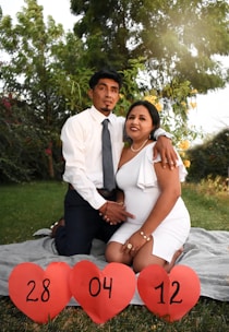 A couple is sitting on a blanket in a garden setting. The woman is wearing a white dress and the man is wearing a white shirt with a tie. They are holding hands and smiling. In front of them, three red heart-shaped signs display the numbers 28, 04, and 12.
