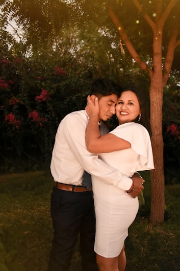 A couple is embracing under a tree in a garden setting, with warm, golden light filtering through the branches. The woman is smiling at the camera while the man holds her close with his eyes closed.