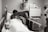 A person lying in a hospital bed receiving a kiss on the forehead from another person leaning over. The room contains medical equipment, including an IV drip and various monitors on the wall. The setting appears to be a hospital or medical facility.