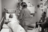 A hospital room with a patient lying in a bed, covered with a blanket and wearing protective eyewear. A medical professional in scrubs and a cap is standing beside the bed, possibly speaking to the patient or performing a task. Another person is seated on a chair nearby, observing the scene.