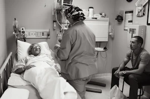 A hospital room with a patient lying in a bed, covered with a blanket and wearing protective eyewear. A medical professional in scrubs and a cap is standing beside the bed, possibly speaking to the patient or performing a task. Another person is seated on a chair nearby, observing the scene.