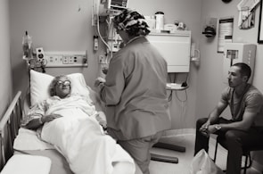 A hospital room with a patient lying in a bed, covered with a blanket and wearing protective eyewear. A medical professional in scrubs and a cap is standing beside the bed, possibly speaking to the patient or performing a task. Another person is seated on a chair nearby, observing the scene.