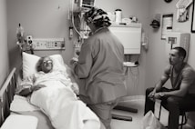 A hospital room with a patient lying in a bed, covered with a blanket and wearing protective eyewear. A medical professional in scrubs and a cap is standing beside the bed, possibly speaking to the patient or performing a task. Another person is seated on a chair nearby, observing the scene.