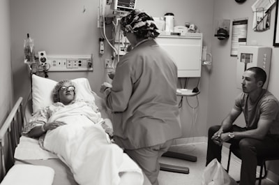A hospital room with a patient lying in a bed, covered with a blanket and wearing protective eyewear. A medical professional in scrubs and a cap is standing beside the bed, possibly speaking to the patient or performing a task. Another person is seated on a chair nearby, observing the scene.