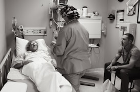 A hospital room with a patient lying in a bed, covered with a blanket and wearing protective eyewear. A medical professional in scrubs and a cap is standing beside the bed, possibly speaking to the patient or performing a task. Another person is seated on a chair nearby, observing the scene.