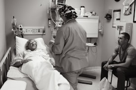 A hospital room with a patient lying in a bed, covered with a blanket and wearing protective eyewear. A medical professional in scrubs and a cap is standing beside the bed, possibly speaking to the patient or performing a task. Another person is seated on a chair nearby, observing the scene.