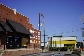 A red brick building with a large sign reading 'Mother Road Market' and a Route 66 emblem on it. The building has black awnings and a railing leading to the entrance. Adjacent is a modern structure with a white facade and a yellow stripe. Overhead, utility poles and wires stretch across a clear blue sky.