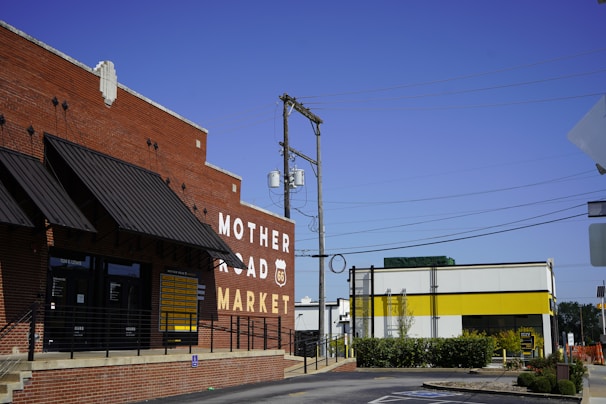 A red brick building with a large sign reading 'Mother Road Market' and a Route 66 emblem on it. The building has black awnings and a railing leading to the entrance. Adjacent is a modern structure with a white facade and a yellow stripe. Overhead, utility poles and wires stretch across a clear blue sky.