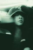 A person wearing a hat with their eyes closed, appearing relaxed or contemplative. The image has a soft, green monochrome effect, creating a calm and serene atmosphere. The word 'EXISTENCE' is visible on the person's clothing.