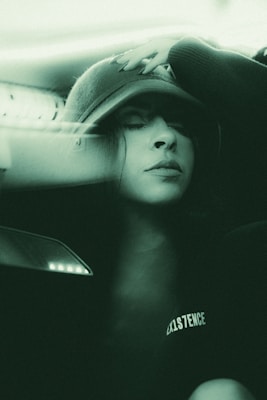 A person wearing a hat with their eyes closed, appearing relaxed or contemplative. The image has a soft, green monochrome effect, creating a calm and serene atmosphere. The word 'EXISTENCE' is visible on the person's clothing.
