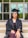 A person wearing a graduation cap and gown is sitting on steps with hands placed on their lap. The background shows a building entrance with glass doors and windows.