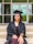 A person wearing a graduation cap and gown is sitting on steps with hands placed on their lap. The background shows a building entrance with glass doors and windows.