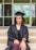 A person wearing a graduation cap and gown is sitting on steps with hands placed on their lap. The background shows a building entrance with glass doors and windows.