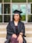 A person wearing a graduation cap and gown is sitting on steps with hands placed on their lap. The background shows a building entrance with glass doors and windows.