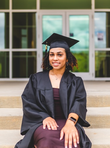 A person wearing a graduation cap and gown is sitting on steps with hands placed on their lap. The background shows a building entrance with glass doors and windows.