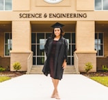 A person wearing a graduation gown and cap stands confidently in front of a building labeled 'Science & Engineering'. The architecture features brickwork and large windows, with a well-maintained pathway and small shrubs leading to the entrance.
